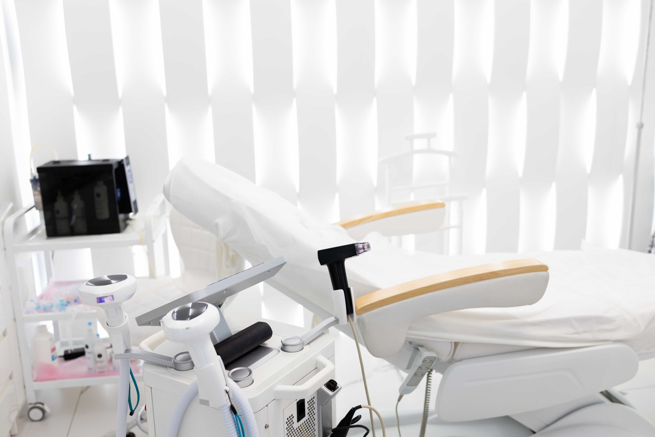 Clean, modern aesthetic treatment room featuring a reclining bed, white privacy blinds, and several pieces of advanced cosmetic technology equipment.