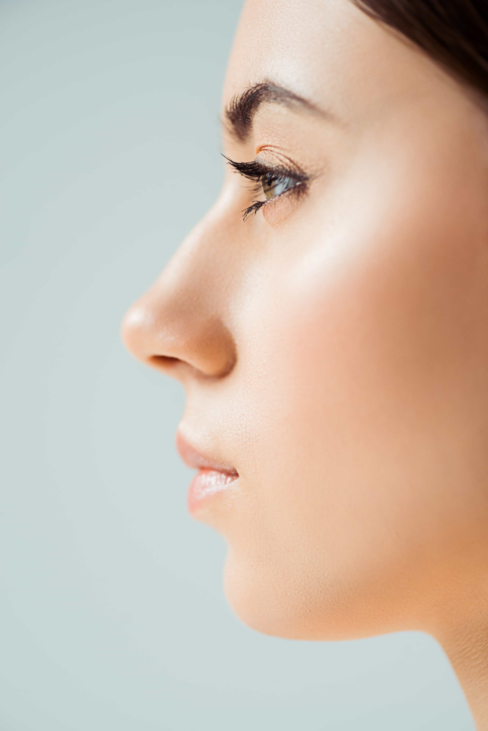Close-up side profile of a woman's face showing a straight nose bridge, full lips, and a defined chin, with a soft blue background.