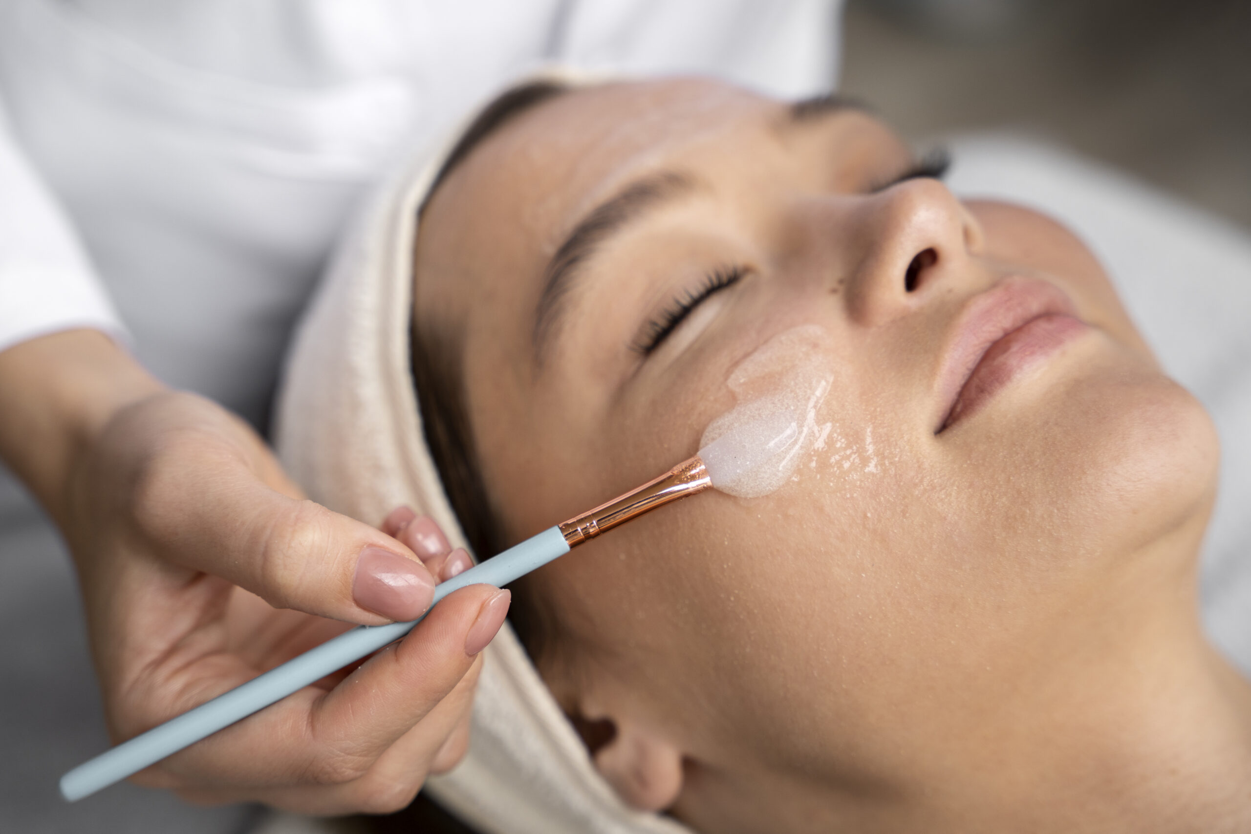 Close-up of a woman receiving a facial treatment, with a clear exfoliating or moisturizing mask being applied to her cheek using a small brush.