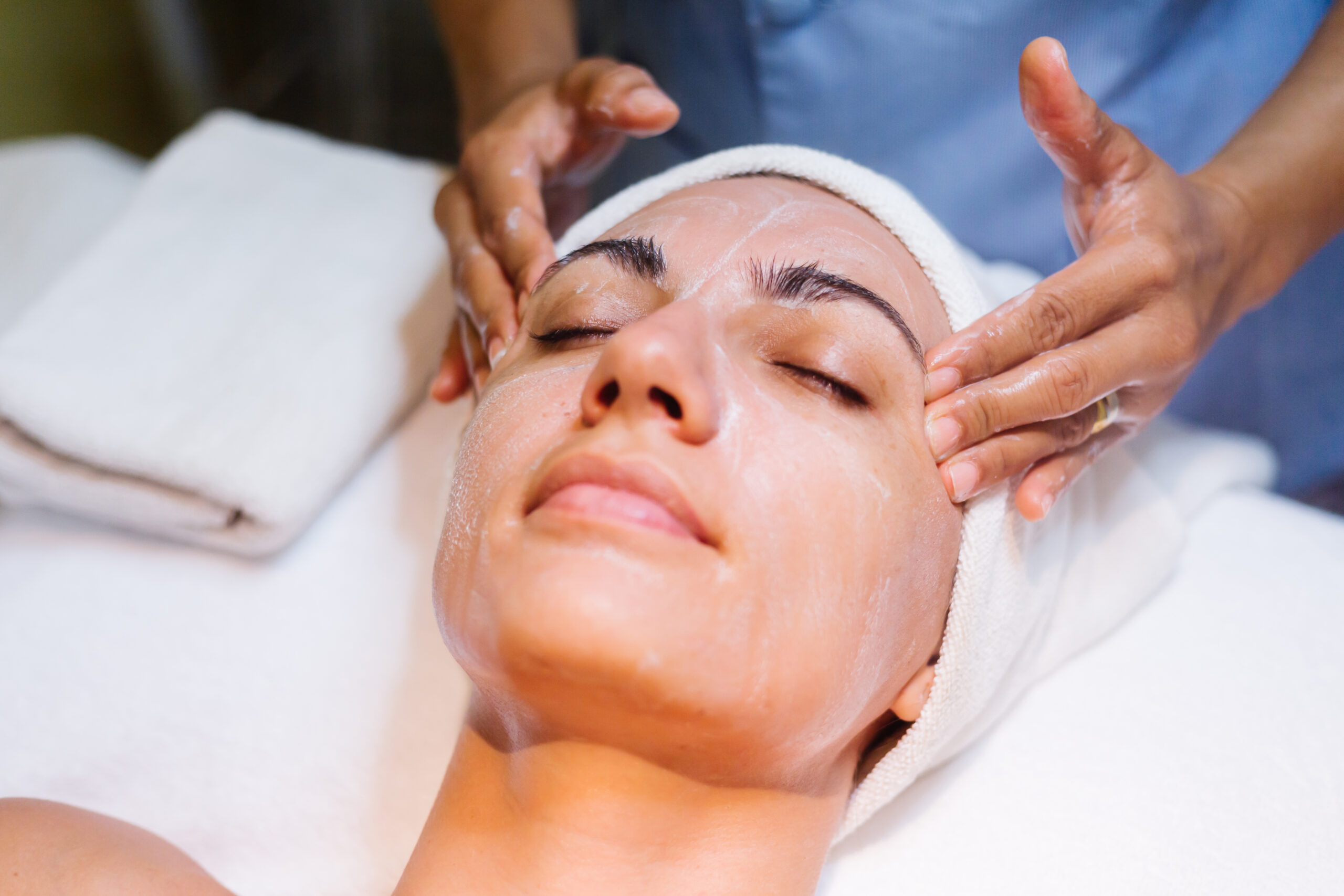 Alt Text: Woman lying down with her eyes closed, receiving a soothing facial massage with a cream or mask being applied by an aesthetician.