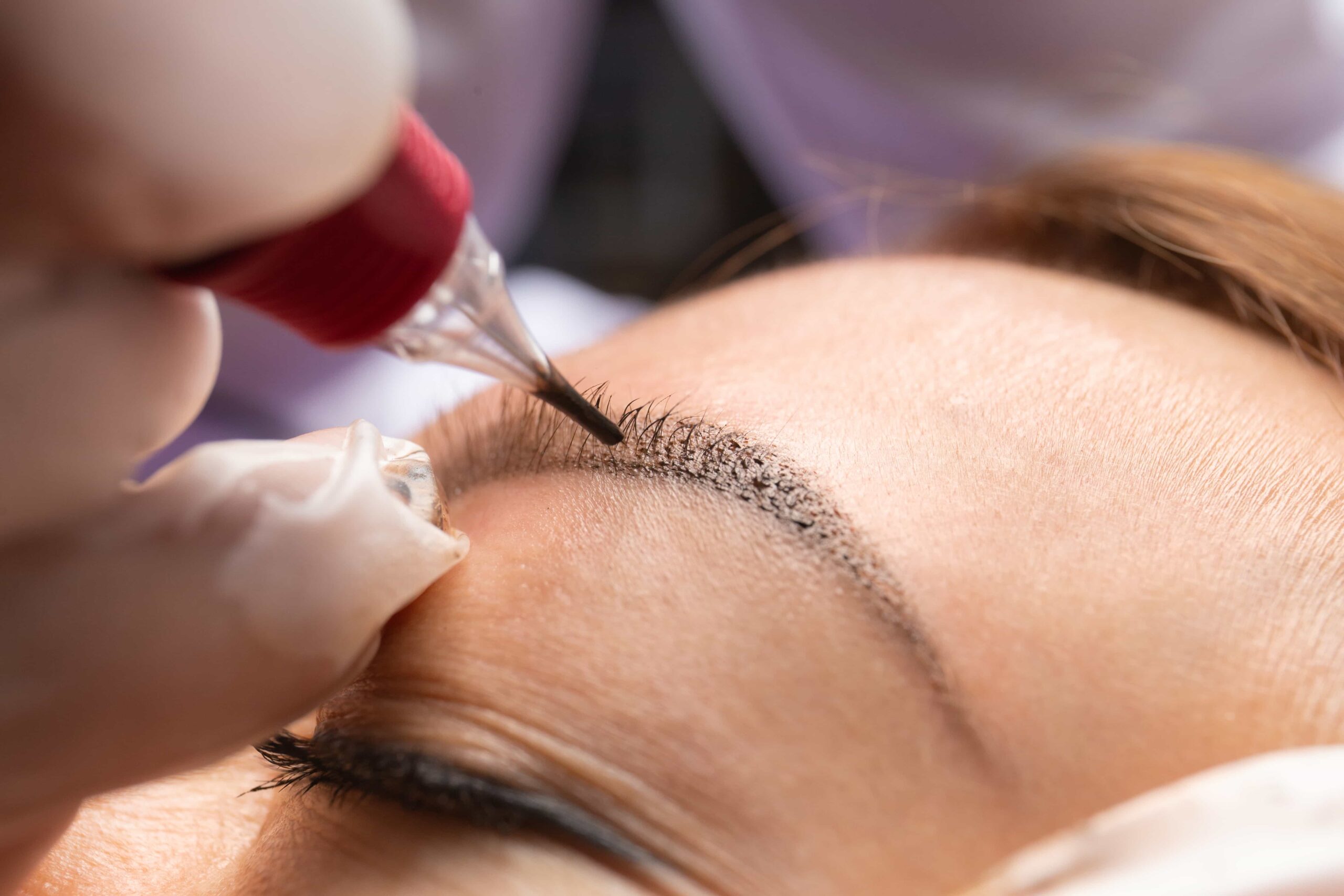Close-up of a professional in white gloves using a permanent makeup device to create natural-looking strokes for an eyebrow microblading or tattooing procedure.