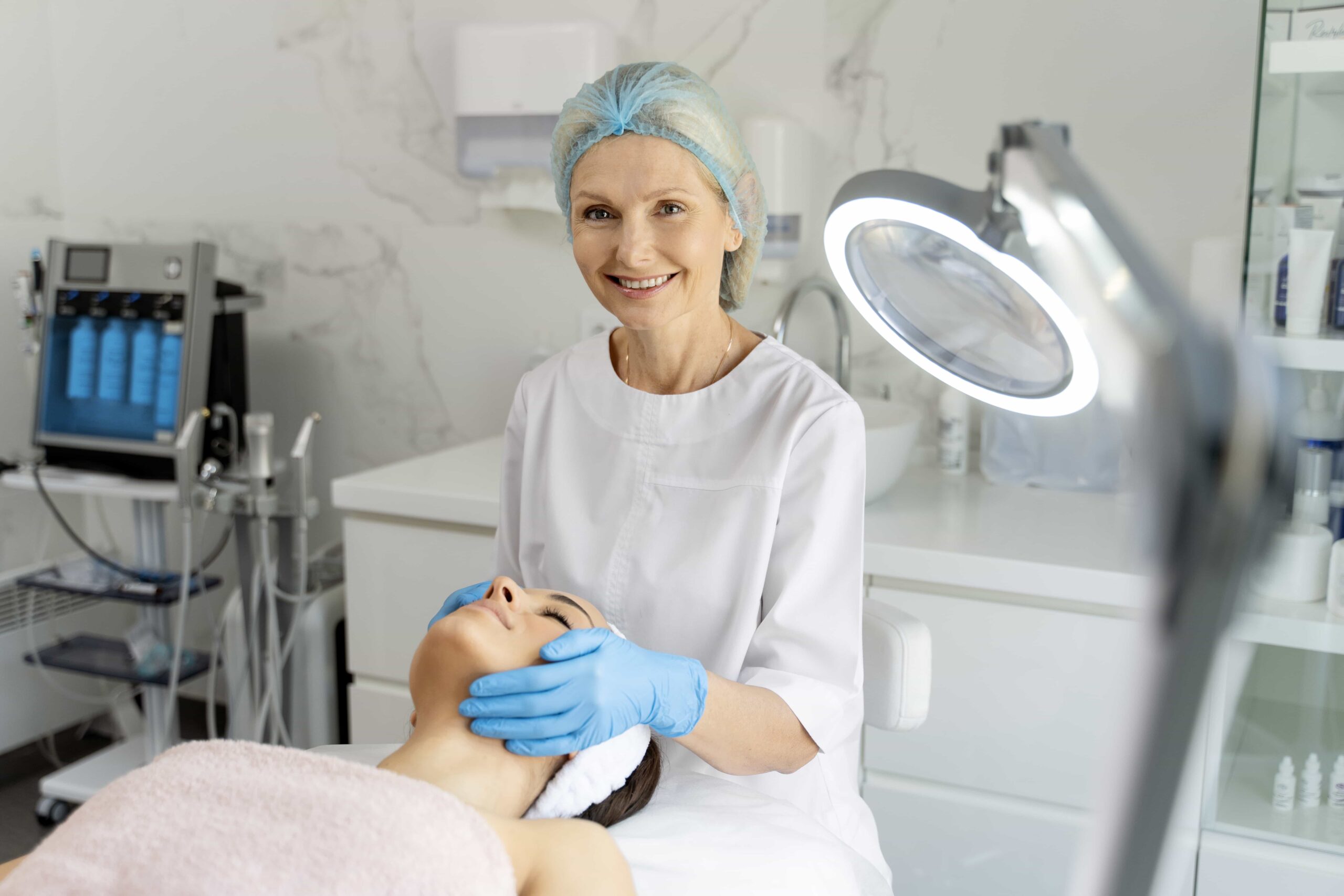 Smiling senior aesthetician in a white uniform and blue gloves examining a younger client's face under a magnifying lamp in a bright clinic.